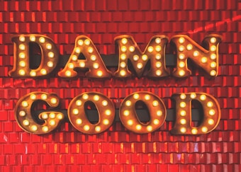 Large, illuminated letters spell out 'DAMN GOOD' against a textured, bright red background. Each letter is outlined with small, retro-style light bulbs that add a vibrant glow to the composition.