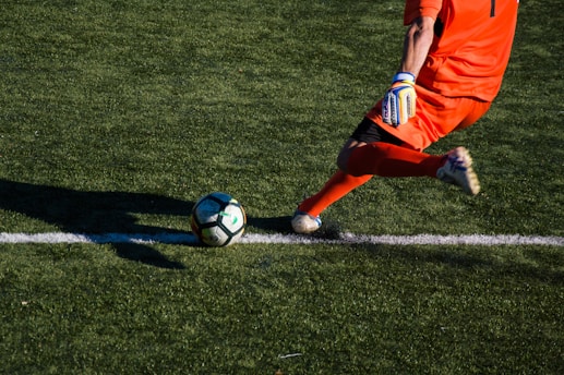 A soccer player wearing bright orange kit and gloves is in the process of kicking a soccer ball on a grass field. The sunlight casts a clear shadow of the player on the ground, adding a dynamic feel to the scene.