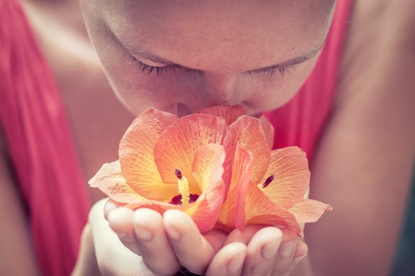 A person with their eyes closed is holding and deeply inhaling the fragrance of an orange hibiscus flower with both hands cupped underneath. The soft focus background adds a calming effect, highlighting the serene moment.