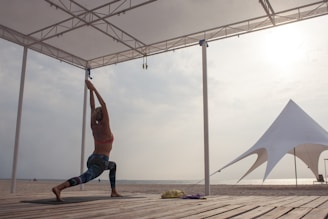A person is practicing yoga in a warrior pose on a wooden platform by the beach, under a canopy structure. The background includes a large white tent and the sun is shining through a cloudy sky.