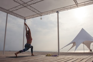 A person is practicing yoga in a warrior pose on a wooden platform by the beach, under a canopy structure. The background includes a large white tent and the sun is shining through a cloudy sky.