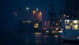 A large cargo ship is seen docked at a port during nighttime, with bright lights illuminating its structure. Other smaller boats and industrial machinery are visible in the background, partially obscured by shadows. The water reflects the lights, adding a shimmering effect to the scene.