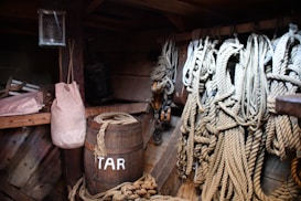 A collection of nautical ropes hangs neatly from hooks on a wooden wall. Nearby, a wooden barrel labeled 'TAR' and other maritime tools and supplies are visible. There is a cloth bag hanging and some rolls of rope on a shelf in an enclosed, wooden ship's storage area.