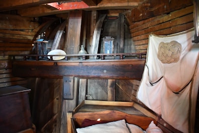 An interior setting with wooden walls and beams, displaying a rustic and cozy atmosphere. There is a wooden shelf holding various items including a lantern, a bottle, and a decorative object. A wooden bed with cushions is visible, along with a hanging fabric organizer featuring a rope design.