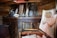 An interior setting with wooden walls and beams, displaying a rustic and cozy atmosphere. There is a wooden shelf holding various items including a lantern, a bottle, and a decorative object. A wooden bed with cushions is visible, along with a hanging fabric organizer featuring a rope design.