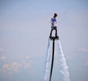 A person is riding a flyboard, elevated above the water with jets of water propelling them into the sky. The individual is wearing shorts and a long-sleeve top, with a serene blue sky as the background.