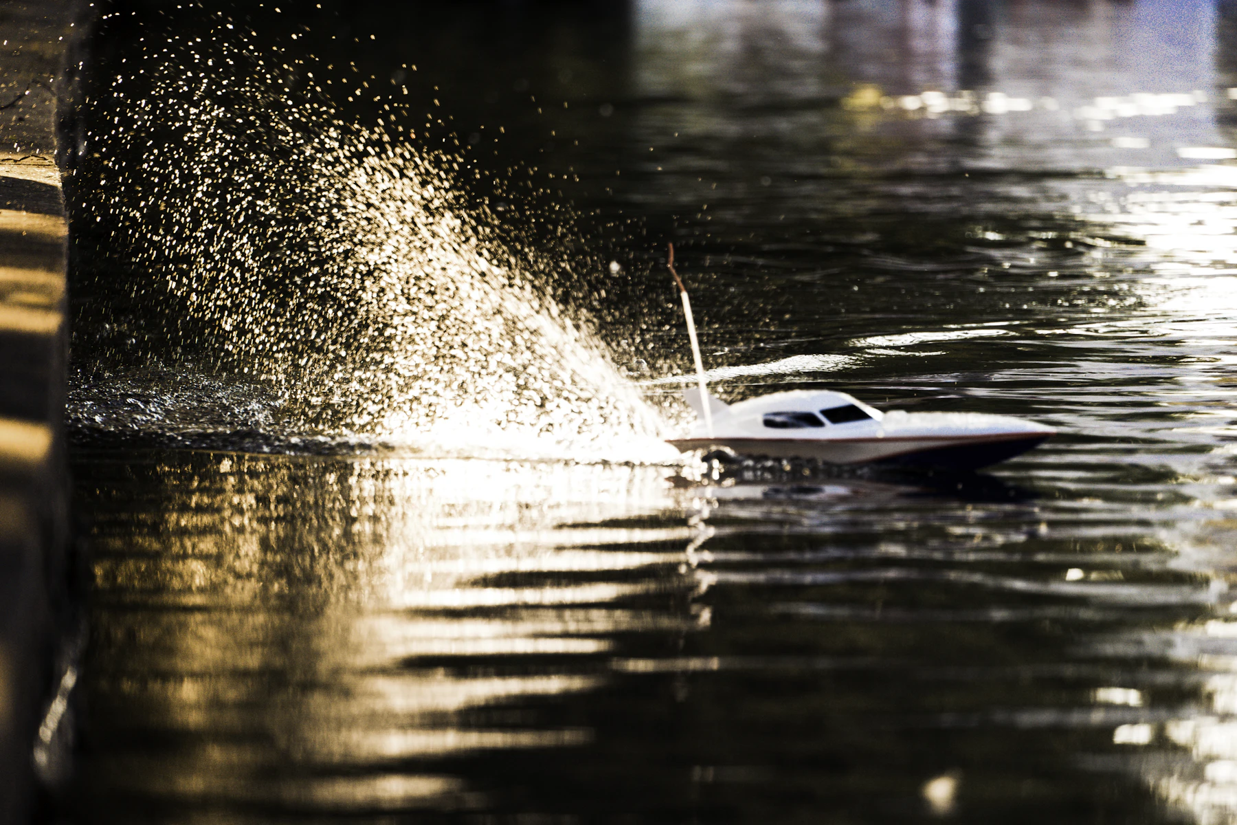 A white radio control speed boat with a whip antenna throwing a backlit plume of spray across dark water