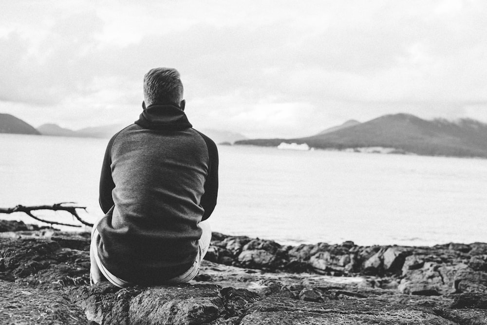 man in black hoodie sitting on rock near body of water during daytime