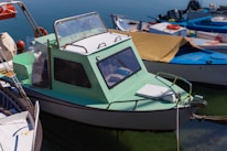 A small, mint-green motorboat with a cabin docked among other boats in a marina, surrounded by calm, clear water. The boat has a simple design with windows on the cabin and a white hatch on the deck.