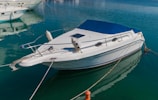 A sleek white motorboat is moored in calm, crystal-clear water. The boat features a blue canopy and metal railings, with mooring lines securing it to the dock. Other boats are visible in the background, enhancing the tranquil marina setting.
