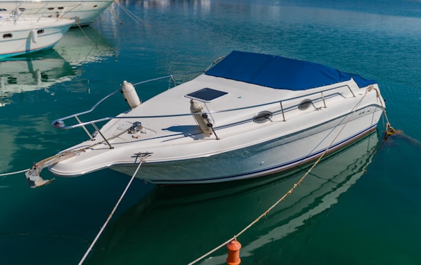 A sleek white motorboat is moored in calm, crystal-clear water. The boat features a blue canopy and metal railings, with mooring lines securing it to the dock. Other boats are visible in the background, enhancing the tranquil marina setting.
