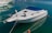A sleek white motorboat is moored in calm, crystal-clear water. The boat features a blue canopy and metal railings, with mooring lines securing it to the dock. Other boats are visible in the background, enhancing the tranquil marina setting.