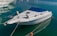 A sleek white motorboat is moored in calm, crystal-clear water. The boat features a blue canopy and metal railings, with mooring lines securing it to the dock. Other boats are visible in the background, enhancing the tranquil marina setting.