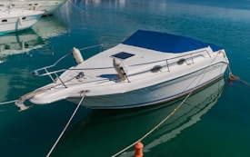 A sleek white motorboat is moored in calm, crystal-clear water. The boat features a blue canopy and metal railings, with mooring lines securing it to the dock. Other boats are visible in the background, enhancing the tranquil marina setting.