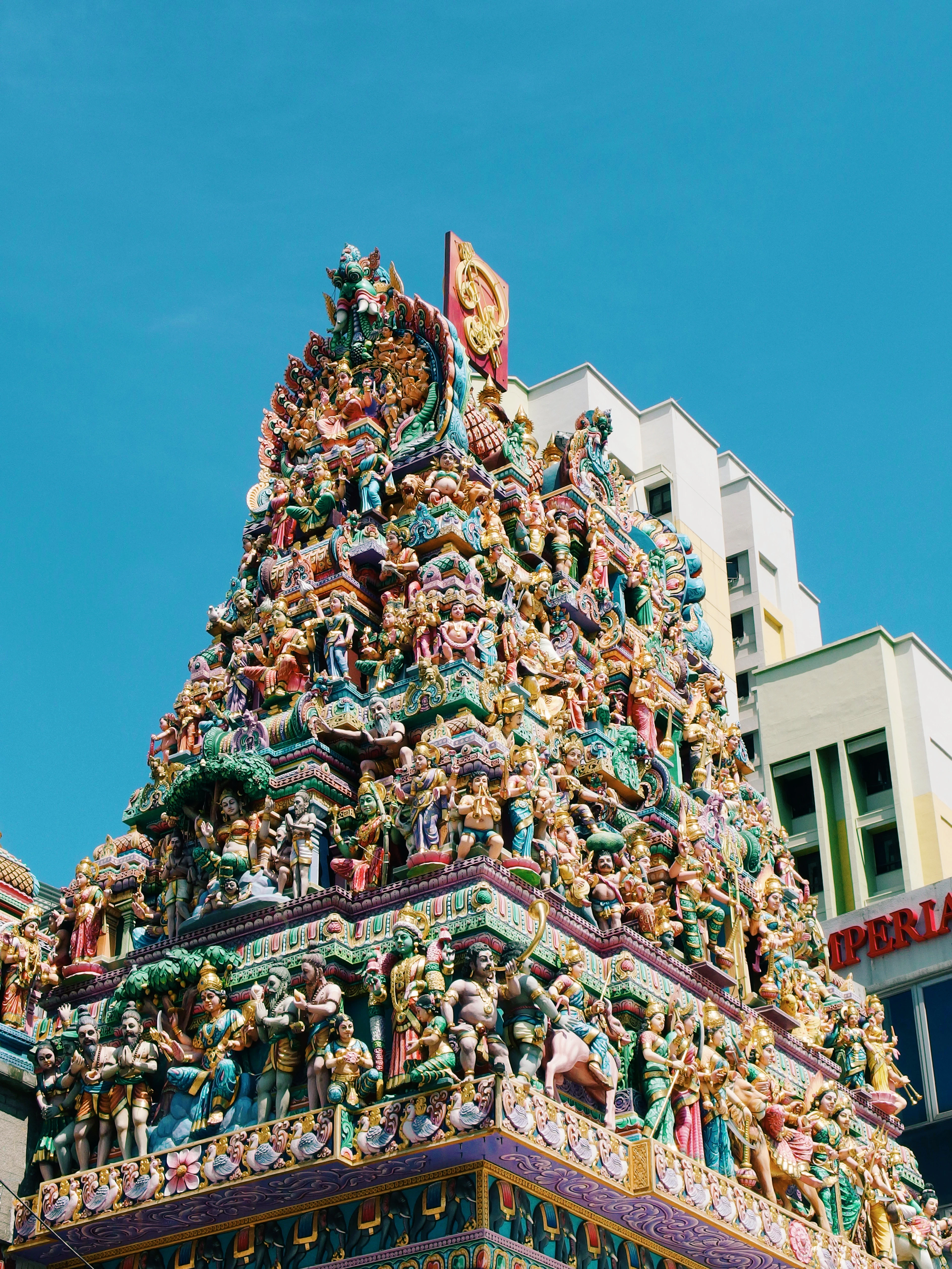 The gopuram tower of the Sri Veeramakaliamman Temple in Singapore's Little India, densely packed with brightly painted figures of Hindu deities, rising against a clear deep blue sky.