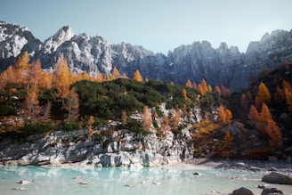 A serene landscape featuring a turquoise lake in the foreground with rocky terrain around it. The middle ground is filled with lush greenery interspersed with tall, orange fall trees, leading up to a range of rugged, gray mountains in the background under a pale blue sky.