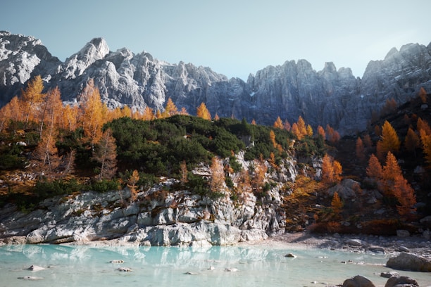 A serene landscape featuring a turquoise lake in the foreground with rocky terrain around it. The middle ground is filled with lush greenery interspersed with tall, orange fall trees, leading up to a range of rugged, gray mountains in the background under a pale blue sky.