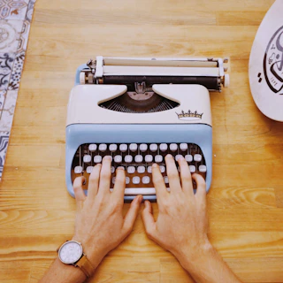 Close-up of a hand typing on a vintage typewriter with sinkhole quarterly cover pages scattered nearby.