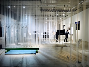 An art exhibition space with transparent curtains hanging from the ceiling, creating a layered and open layout. The room is well-lit with spotlights on the ceiling. People are walking and interacting with various installations and displays, some of which consist of framed artworks and digital screens. The concrete floor and minimalistic design contribute to a modern gallery atmosphere.