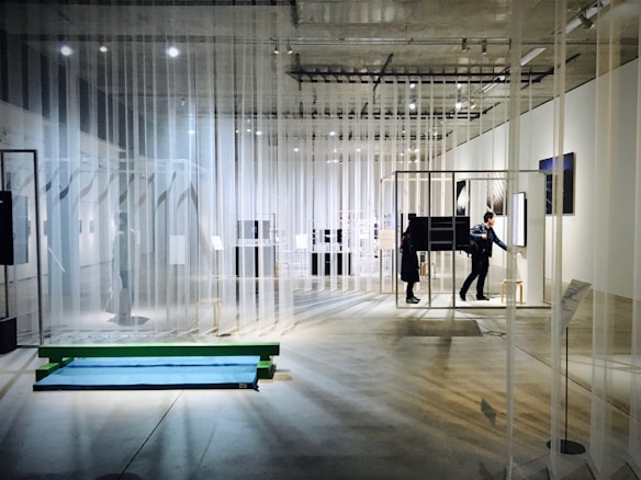 An art exhibition space with transparent curtains hanging from the ceiling, creating a layered and open layout. The room is well-lit with spotlights on the ceiling. People are walking and interacting with various installations and displays, some of which consist of framed artworks and digital screens. The concrete floor and minimalistic design contribute to a modern gallery atmosphere.
