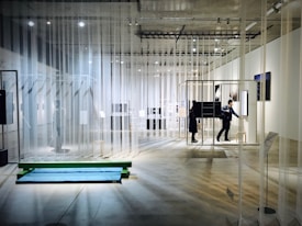 An art exhibition space with transparent curtains hanging from the ceiling, creating a layered and open layout. The room is well-lit with spotlights on the ceiling. People are walking and interacting with various installations and displays, some of which consist of framed artworks and digital screens. The concrete floor and minimalistic design contribute to a modern gallery atmosphere.