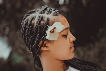 A person with intricately braided hair adorned with a white flower is shown in profile against a softly blurred background, highlighting the texture of the braids and the delicate petals of the flower.