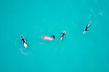 Four surfers wearing wetsuits float on surfboards in the vibrant turquoise water. They are spaced apart, leisurely lying on their boards, indicating a relaxed and calm scene.