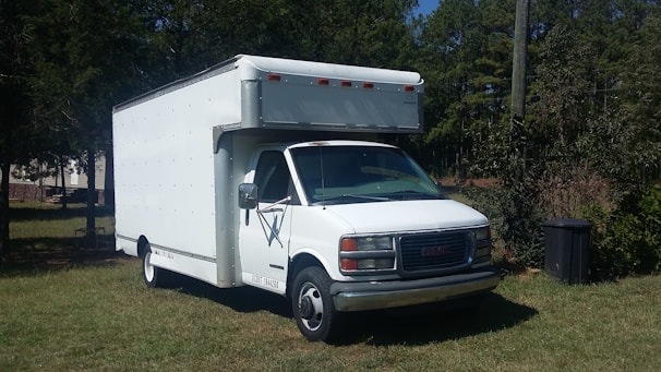 A white box truck with a prominent cab is parked on a grassy area surrounded by trees. The vehicle appears to be in good condition, with a clean exterior and visible side mirrors. The area is rural, with some trees in the background providing greenery.