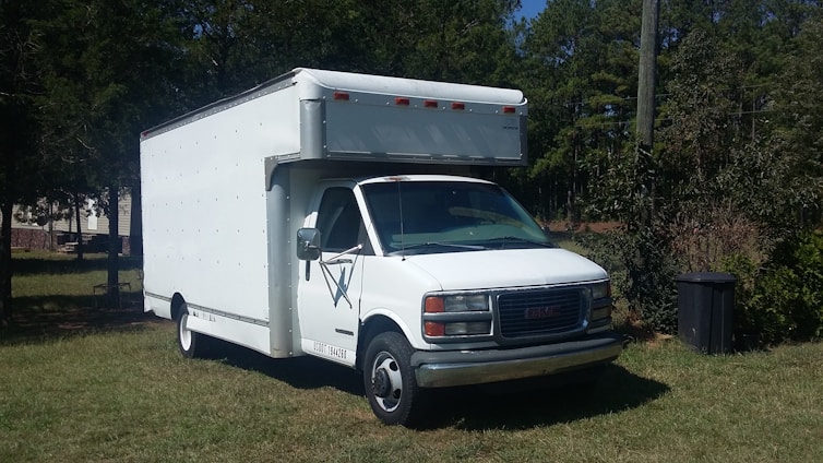 A white box truck with a prominent cab is parked on a grassy area surrounded by trees. The vehicle appears to be in good condition, with a clean exterior and visible side mirrors. The area is rural, with some trees in the background providing greenery.