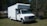 A white box truck with a prominent cab is parked on a grassy area surrounded by trees. The vehicle appears to be in good condition, with a clean exterior and visible side mirrors. The area is rural, with some trees in the background providing greenery.