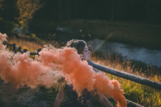 A young person stands outdoors near a wooden fence with an orange smoke bomb billowing around them. The background features a river and grassy area with trees in the distance, creating a tranquil setting.