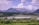 A rural landscape with a series of barns and a small house in the foreground, surrounded by lush green vegetation and fields. Rolling hills and dense pine forests lie behind the buildings, leading up to a range of majestic mountains under a partly cloudy sky.
