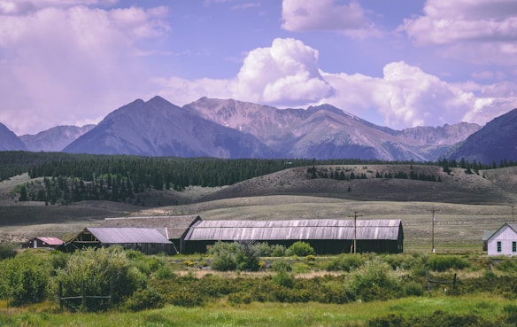 A rural landscape with a series of barns and a small house in the foreground, surrounded by lush green vegetation and fields. Rolling hills and dense pine forests lie behind the buildings, leading up to a range of majestic mountains under a partly cloudy sky.