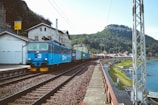 A blue locomotive is pulling a long freight train composed of various colored cargo containers, traveling along railway tracks beside a river. The scene is set in a small town with buildings adjacent to the railway line. In the background, lush green hills are visible, suggesting a rural landscape.