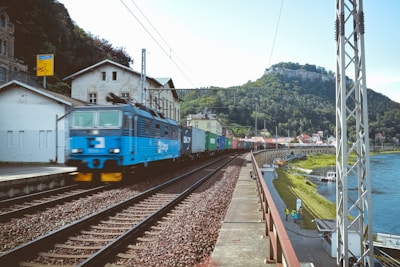 A blue locomotive is pulling a long freight train composed of various colored cargo containers, traveling along railway tracks beside a river. The scene is set in a small town with buildings adjacent to the railway line. In the background, lush green hills are visible, suggesting a rural landscape.