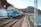 A blue locomotive is pulling a long freight train composed of various colored cargo containers, traveling along railway tracks beside a river. The scene is set in a small town with buildings adjacent to the railway line. In the background, lush green hills are visible, suggesting a rural landscape.