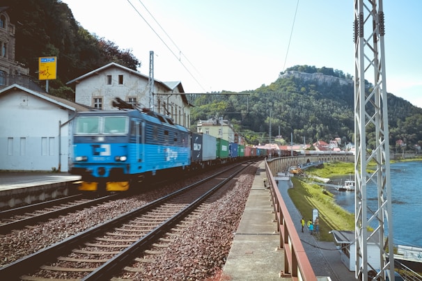 A blue locomotive is pulling a long freight train composed of various colored cargo containers, traveling along railway tracks beside a river. The scene is set in a small town with buildings adjacent to the railway line. In the background, lush green hills are visible, suggesting a rural landscape.