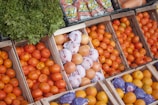 An assortment of fresh produce displayed in wooden crates includes oranges, grapefruits, and leafy greens. Some grapefruits are wrapped in white paper, while others are bare. Crisp vegetables and vibrant fruits are neatly arranged, creating a colorful display. Bags of garlic are visible in the background.