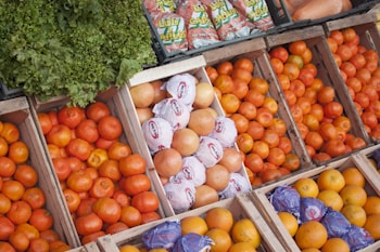 An assortment of fresh produce displayed in wooden crates includes oranges, grapefruits, and leafy greens. Some grapefruits are wrapped in white paper, while others are bare. Crisp vegetables and vibrant fruits are neatly arranged, creating a colorful display. Bags of garlic are visible in the background.