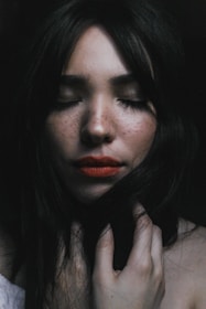 A portrait of a woman with long dark hair and freckles, her eyes closed and her hand gently touching her neck. The mood is serene, with soft lighting creating a calm atmosphere.