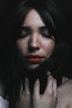 A portrait of a woman with long dark hair and freckles, her eyes closed and her hand gently touching her neck. The mood is serene, with soft lighting creating a calm atmosphere.