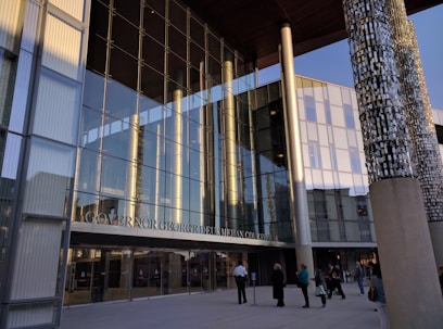 A modern courthouse with large glass windows reflecting the surroundings. The entrance is marked with pillars displaying intricate designs. Several people are walking and gathering near the entrance, wearing formal and casual attire. The evening light casts warm tones on the building.