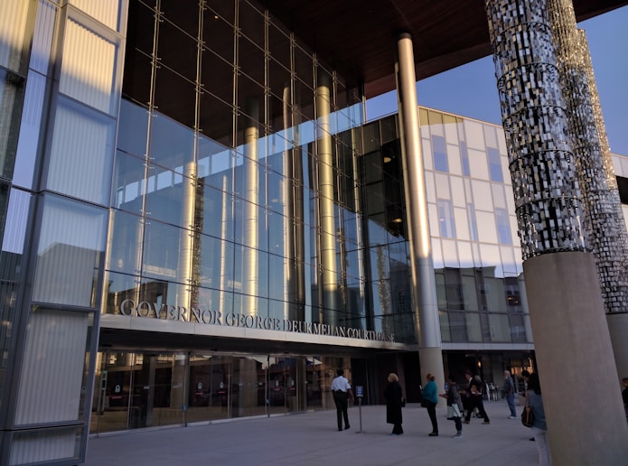 A modern courthouse with large glass windows reflecting the surroundings. The entrance is marked with pillars displaying intricate designs. Several people are walking and gathering near the entrance, wearing formal and casual attire. The evening light casts warm tones on the building.