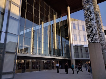 A modern courthouse with large glass windows reflecting the surroundings. The entrance is marked with pillars displaying intricate designs. Several people are walking and gathering near the entrance, wearing formal and casual attire. The evening light casts warm tones on the building.