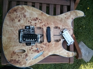 An unfinished electric guitar body is placed on a wooden chair outdoors. The guitar has a natural wood finish with intricate grain patterns. It has a Seymour Duncan pickup installed, and other components are either missing or in the process of being assembled. There is some grass visible on the right side.
