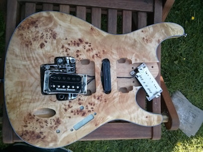 An unfinished electric guitar body is placed on a wooden chair outdoors. The guitar has a natural wood finish with intricate grain patterns. It has a Seymour Duncan pickup installed, and other components are either missing or in the process of being assembled. There is some grass visible on the right side.