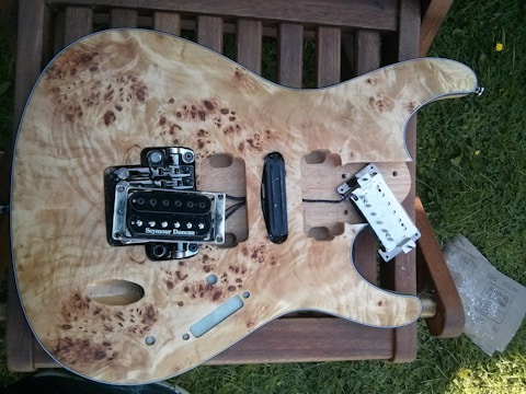 An unfinished electric guitar body is placed on a wooden chair outdoors. The guitar has a natural wood finish with intricate grain patterns. It has a Seymour Duncan pickup installed, and other components are either missing or in the process of being assembled. There is some grass visible on the right side.