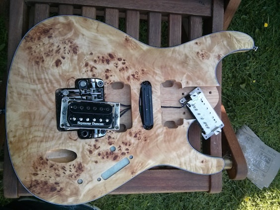 An unfinished electric guitar body is placed on a wooden chair outdoors. The guitar has a natural wood finish with intricate grain patterns. It has a Seymour Duncan pickup installed, and other components are either missing or in the process of being assembled. There is some grass visible on the right side.