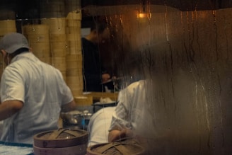 Steamy window partially obscures a busy kitchen where chefs, dressed in white uniforms, are preparing food. Bamboo steamers and stacks of containers are visible. The scene suggests an active environment, likely in a restaurant kitchen during meal preparation.