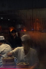 Two individuals are working in a dimly lit kitchen or food preparation area, viewed through a steamy glass. They are dressed in white uniforms and are focused on preparing food using traditional bamboo steamers. The atmosphere is warm and intimate, with reflections of orange and blue lights remaining visible in the glass.